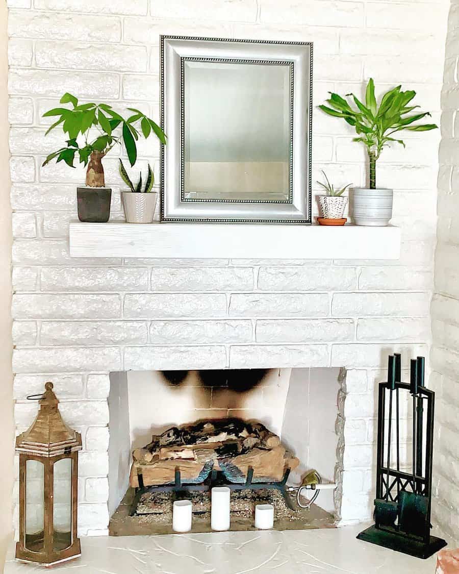 A white brick fireplace with a mantle holding plants and a mirror, three candles, and a lantern sit by the hearth