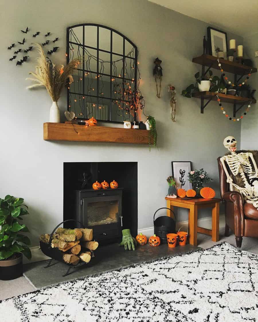 Cozy living room with Halloween decor: skeleton, pumpkins, bats, and orange lights; mirror above mantel, log burner, and patterned rug