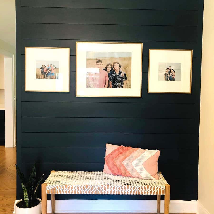 A dark shiplap wall with three framed family photos above a bench featuring a patterned cushion, a pink pillow, and a plant nearby