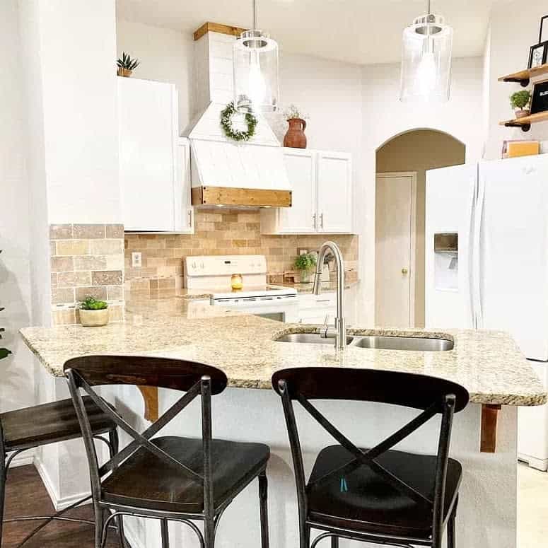 Warm farmhouse kitchen with a granite breakfast bar, black cross-back barstools, white cabinetry, brick backsplash, and glass pendant lighting