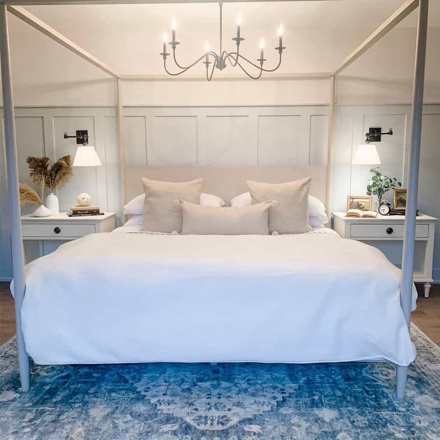 Bedroom with a canopy bed, white linens, and beige pillows; nightstands with lamps and decor on both sides, blue patterned rug underneath