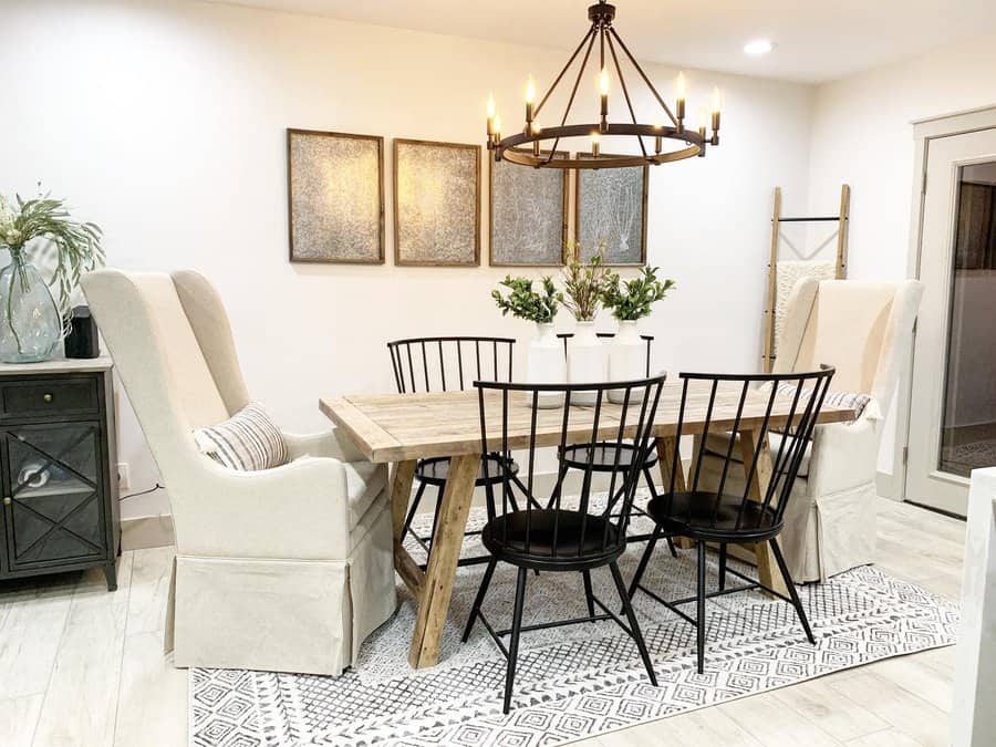 Dining room with a wooden table, black chairs, and cushioned armchairs; a candle-style chandelier hangs above while decor includes plants