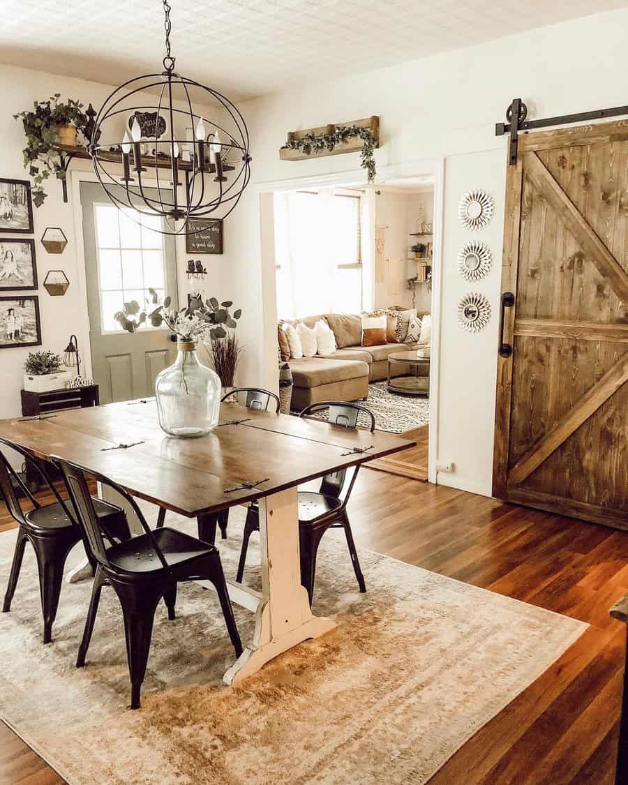 Farmhouse dining room with a wooden table, black chairs, large rustic door, and plants; bright living room is visible in the background