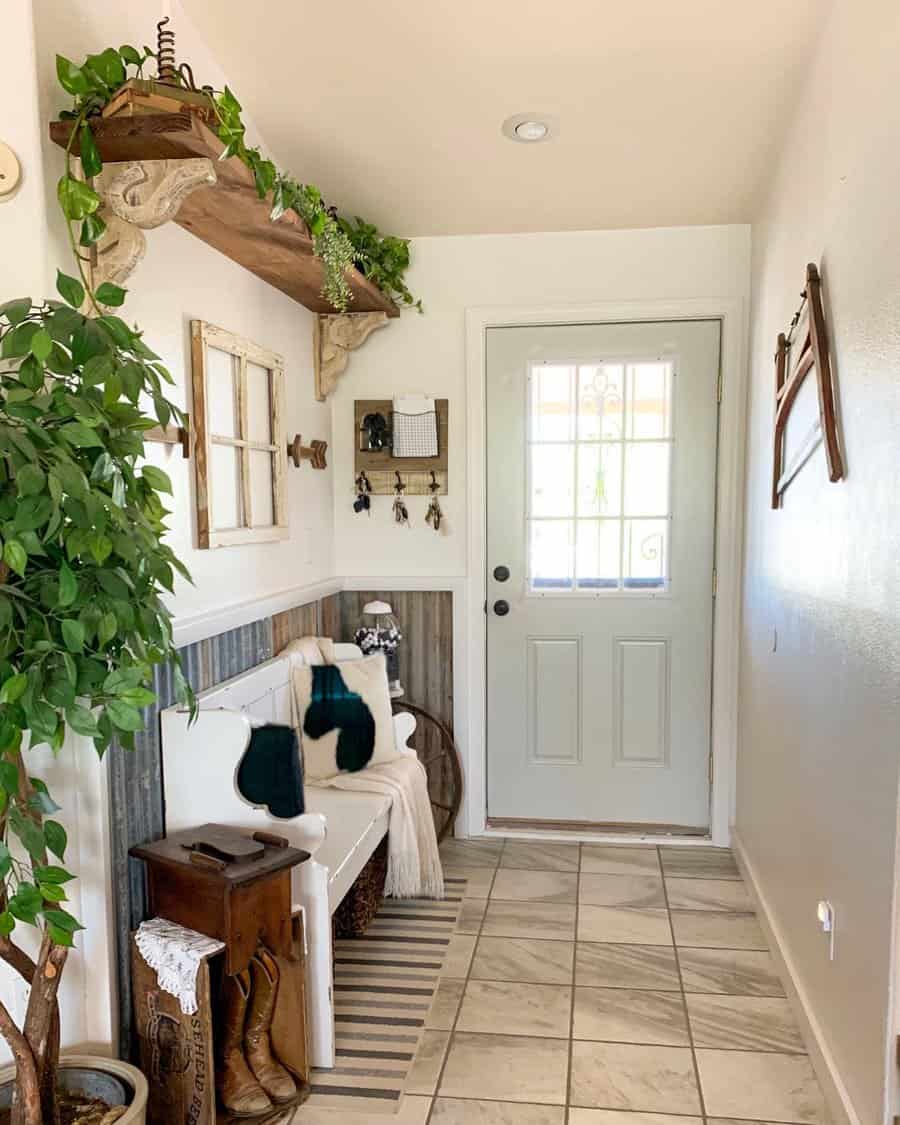 Bright entryway with a bench, boots, greenery, decorative shelf, and wall art; light gray door and tiled floor; cozy and inviting decor