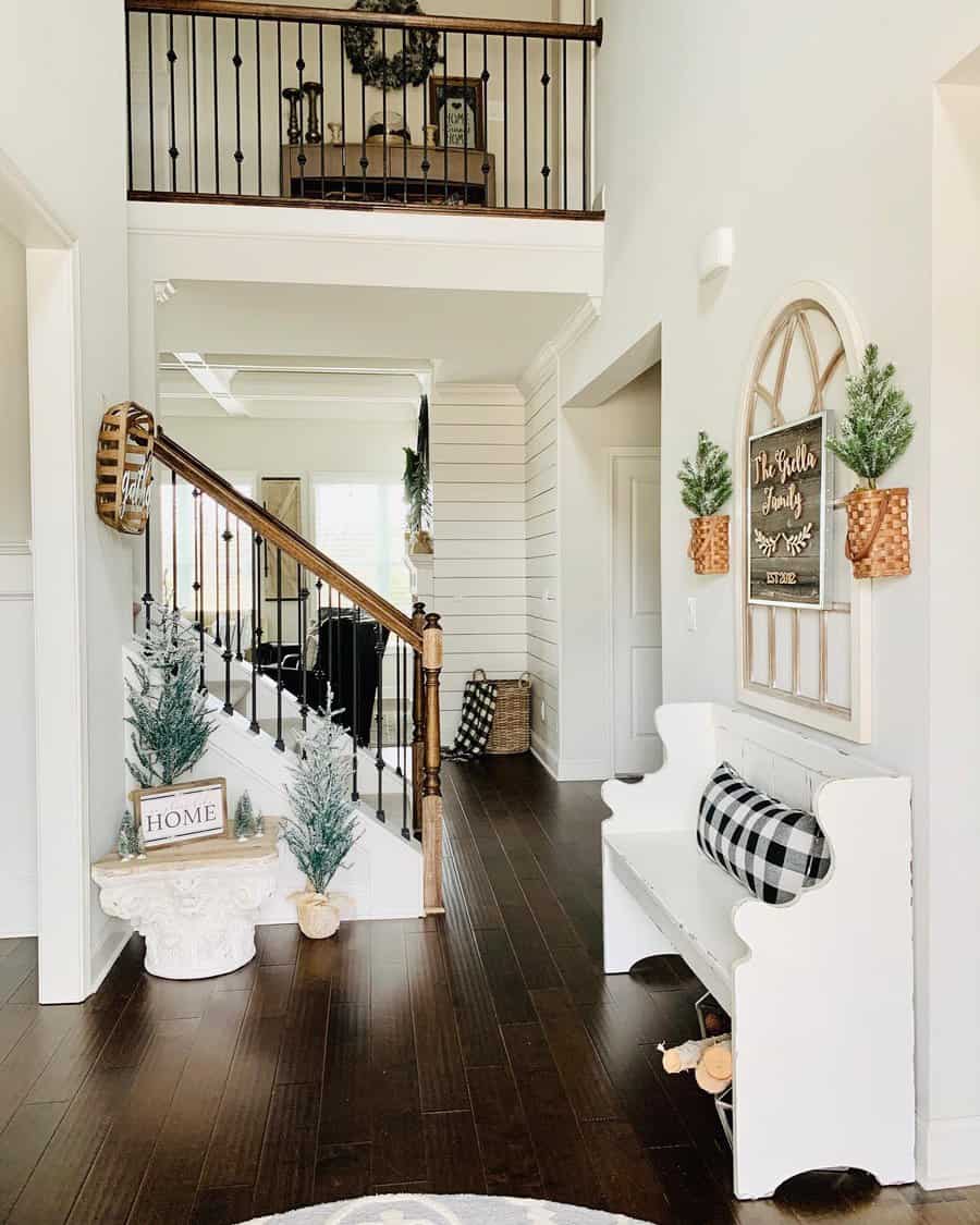 A bright hallway with a white bench, plaid pillows, small trees, and wooden accents featuring stairs with dark rails leading to an upper level