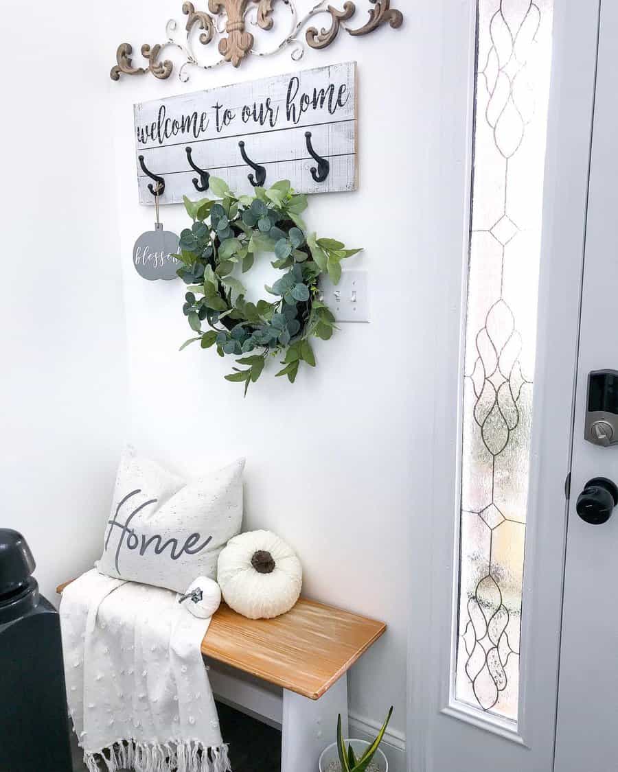 Entryway with a wooden bench, white pillow, blanket, and pumpkin; a wall sign reads "welcome to our home" with a wreath and hooks