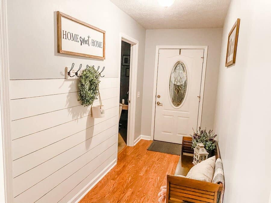 White entryway with wooden bench, shiplap wall, "Home Sweet Home" sign, coat hooks, hanging plant, and oval glass door