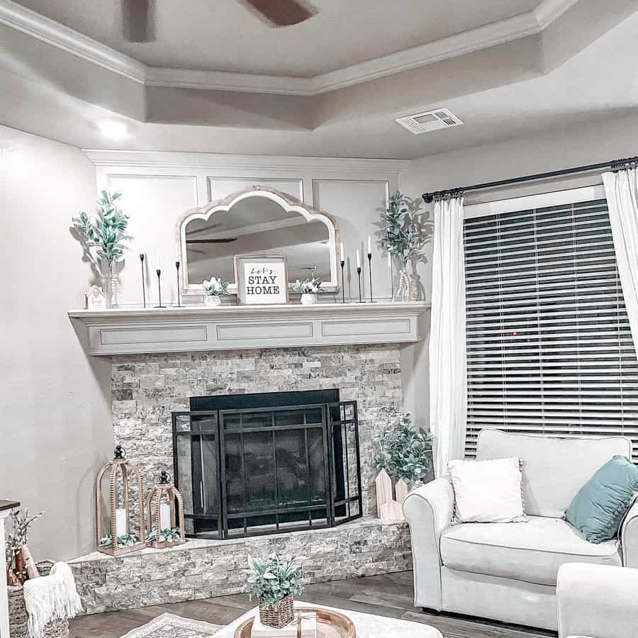 Cozy living room with a fireplace, white sofa, large mirror on the mantel, and decorative plants; bright window with blinds and ceiling fan