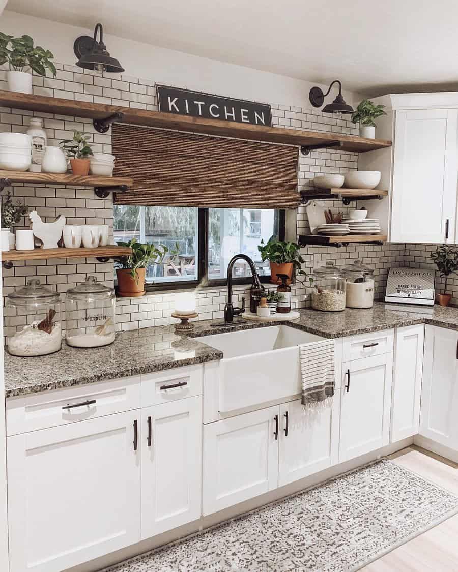 A cozy kitchen with white cabinets, a farmhouse sink, open shelves with plants and dishware, and a window with a rustic shade