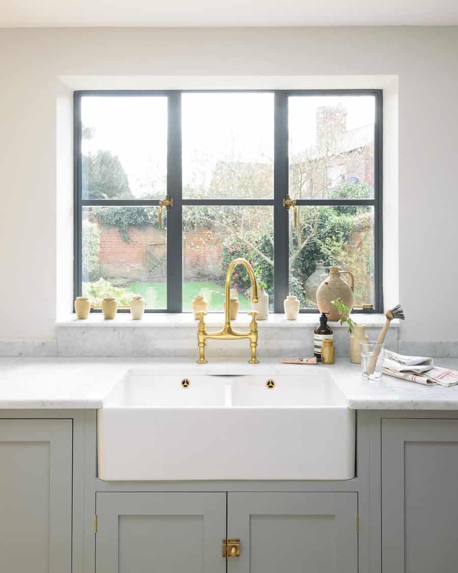 A farmhouse kitchen sink with brass fixtures under a large window, surrounded by plants and ceramic jars