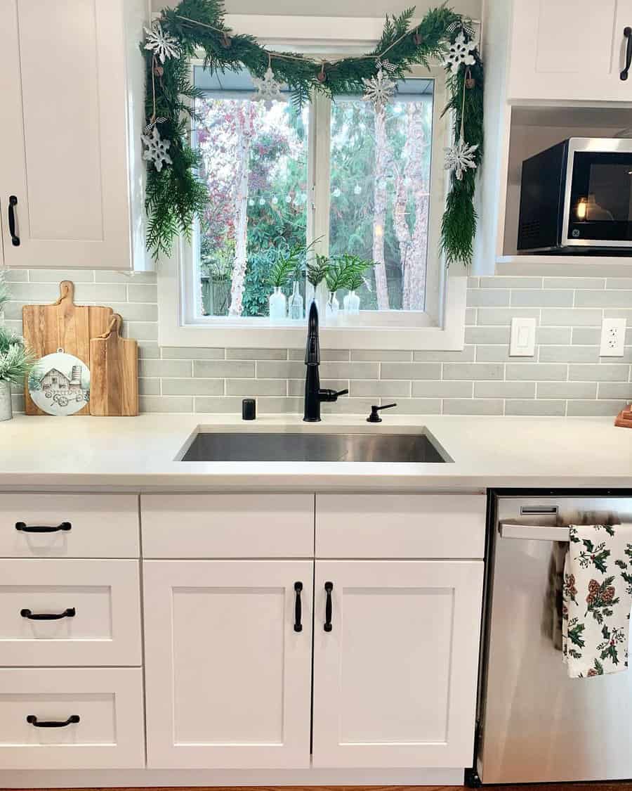 Kitchen sink with black faucet, white cabinets, light green backsplash, and festive garland with snowflakes around the window