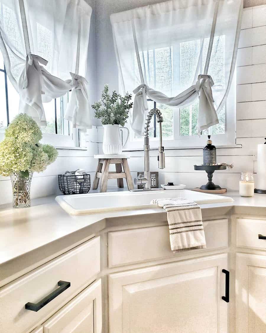 A bright kitchen corner with white cabinets, a farmhouse sink, greenery in a vase, and sheer curtains on two windows