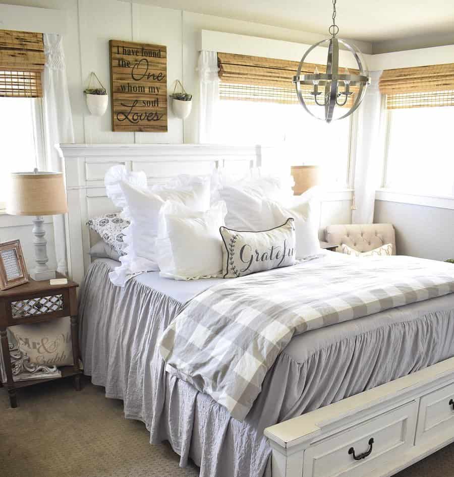 Cozy bedroom with white bed, patterned pillows, rustic decor, and a wooden sign; natural light filters through wicker blinds