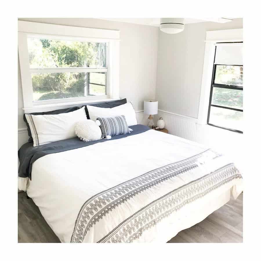 Bright bedroom with a large bed, white linens, decorative pillows, and windows with a view of greenery; lamp on bedside table