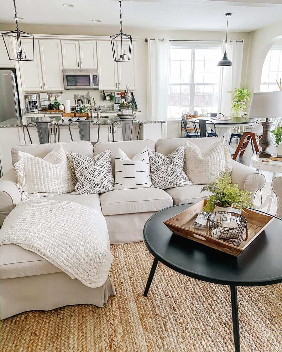 Cozy living room with a light gray sofa, patterned pillows, a knitted throw, black round coffee table, and a modern kitchen in the background