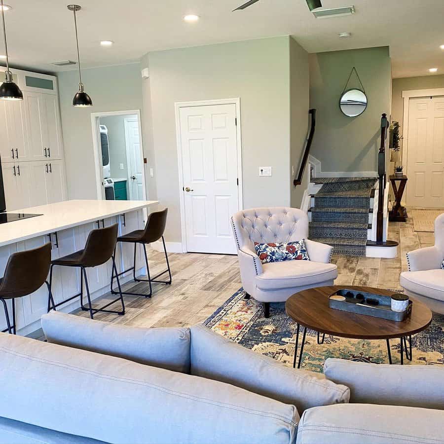 Modern living room with grey sofas, armchairs, a round coffee table, and a kitchen island with bar stools; stairs and mirror in the background