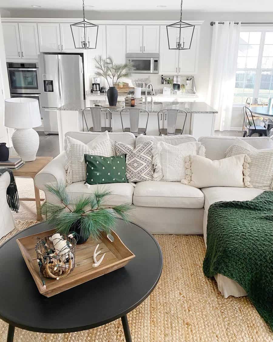 Bright living room with a cozy white sofa, green and patterned pillows, black coffee table, and open kitchen with bar stools in the background