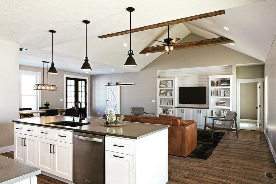 Open-plan living area with a kitchen island, brown sofa, bookshelves, and vaulted ceiling with exposed beams and pendant lights