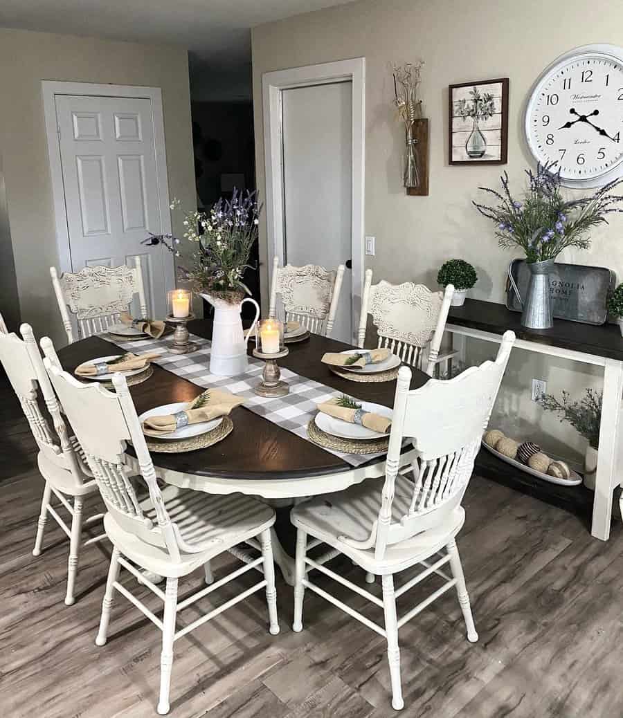 A farmhouse dining room with a wooden table, white chairs, plates, candles, and floral centerpieces, featuring a large wall clock and decor visible