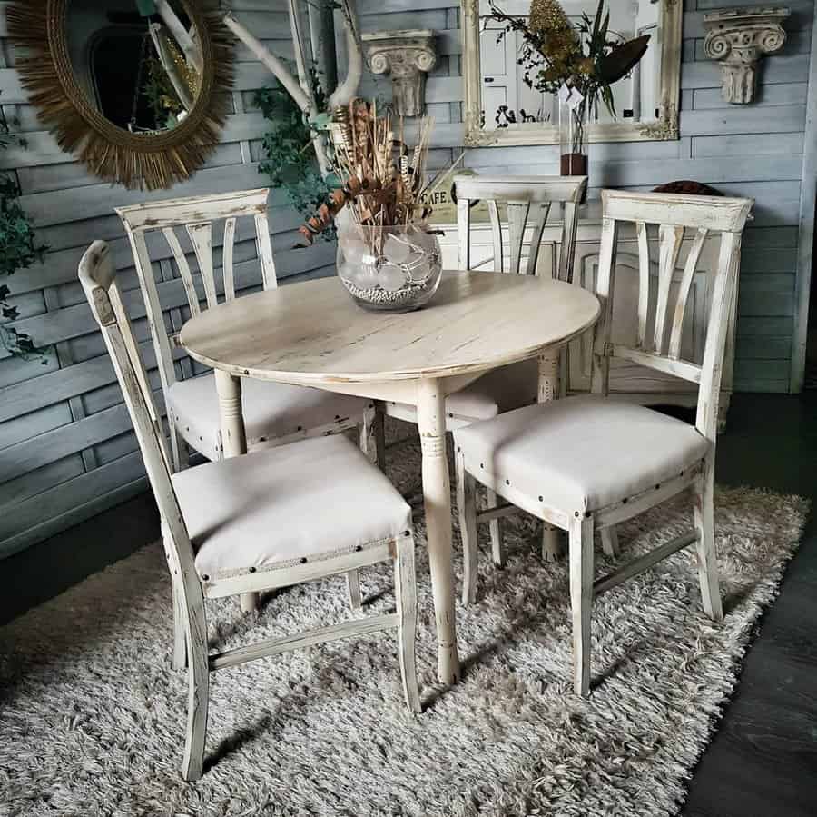 A rustic dining set with a round wooden table and five matching chairs with white cushions on a textured rug in a vintage room