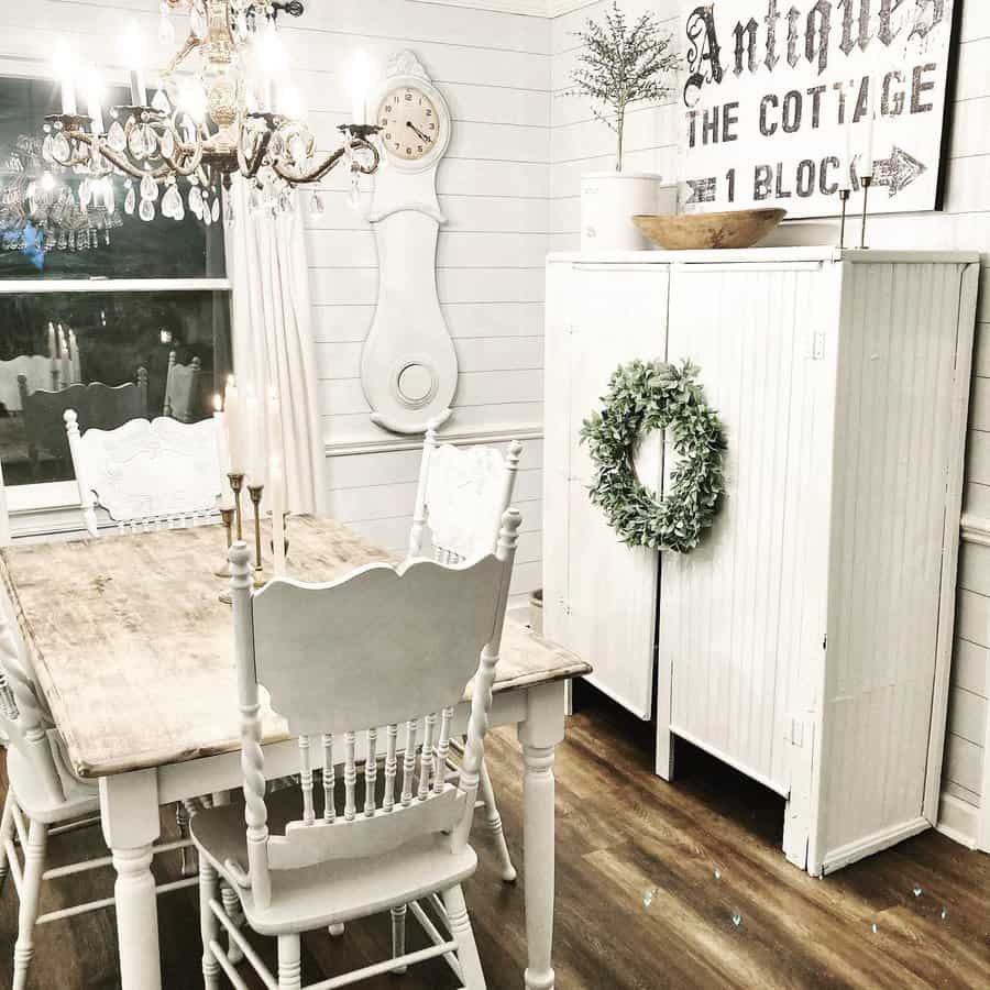 Farmhouse dining room with white vintage furniture, a wooden table, ornate chairs, a chandelier, and a cupboard adorned with a wreath