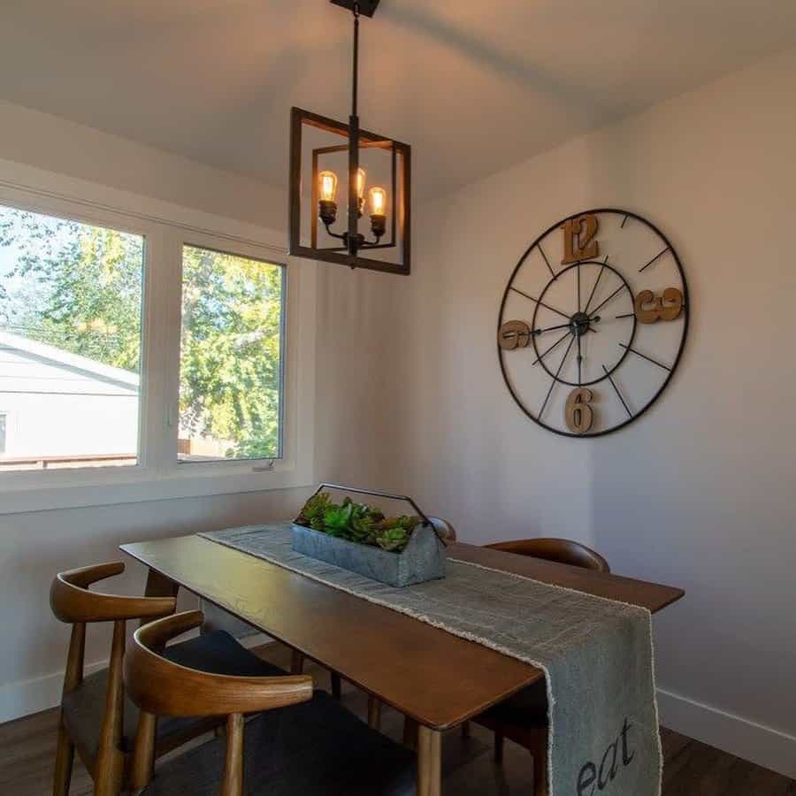 Dining room with wooden table, four chairs, large wall clock, pendant light, and a window showcasing greenery outside