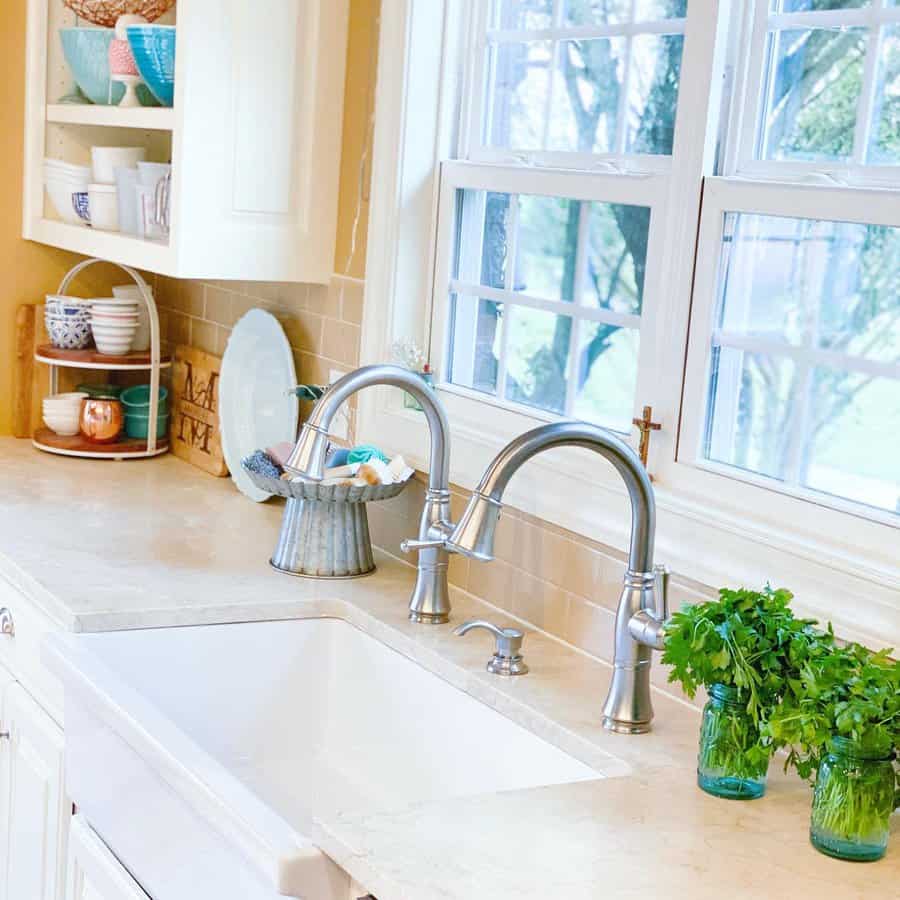 Bright kitchen with a white sink, dual faucets, jars of herbs, and open shelving with dishes, set against large windows