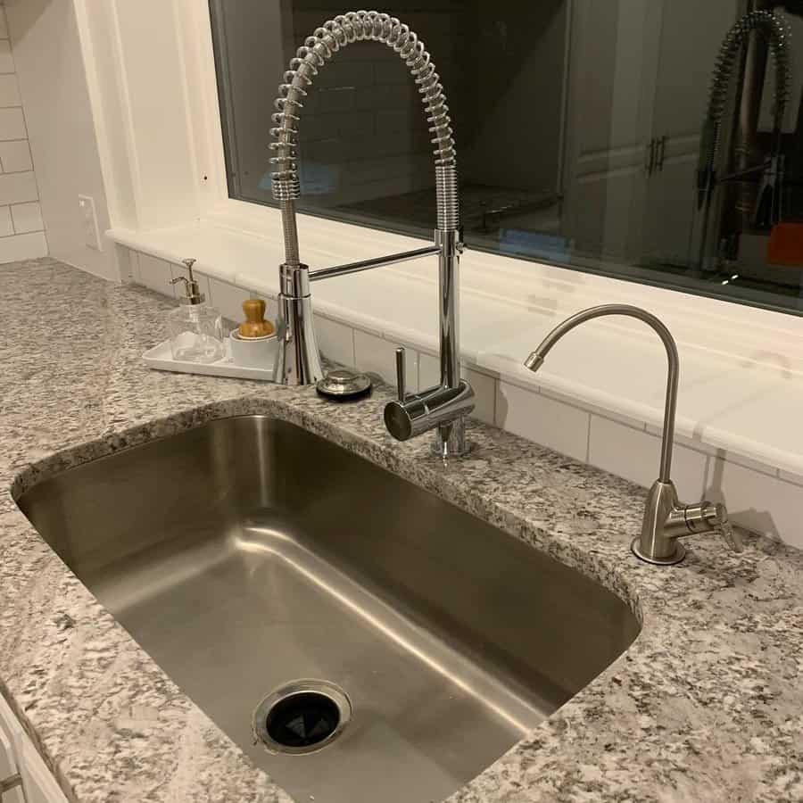 A modern kitchen sink with a tall, coiled faucet and a separate smaller faucet on a granite countertop near a window