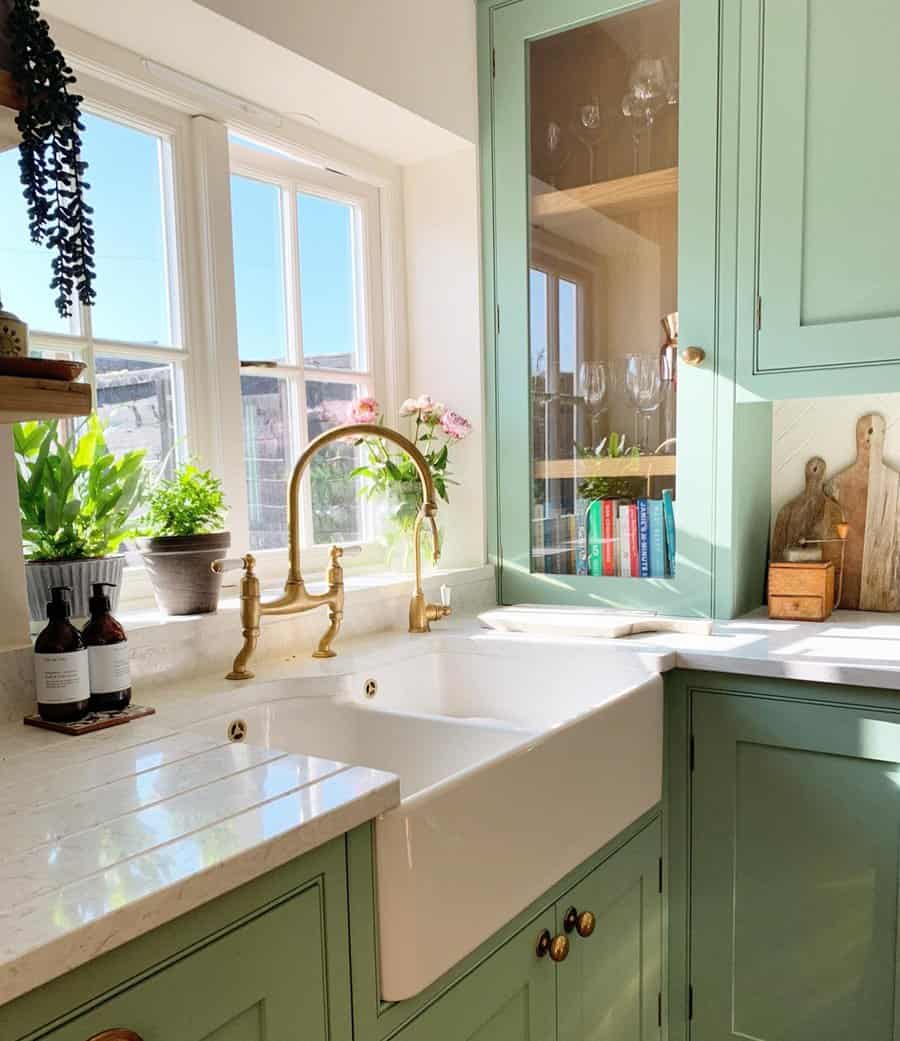 Sunlit kitchen with green cabinets, brass faucet, white farmhouse sink, plants, books, and cutting boards by a window