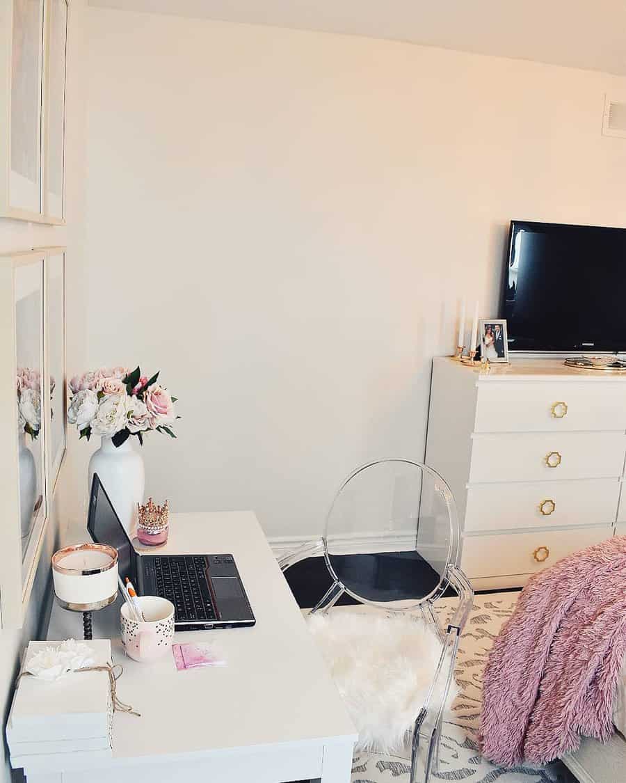 Cozy workspace with a laptop on a white desk, clear chair, flowers in a vase, dresser with a TV, and a bed with a pink blanket