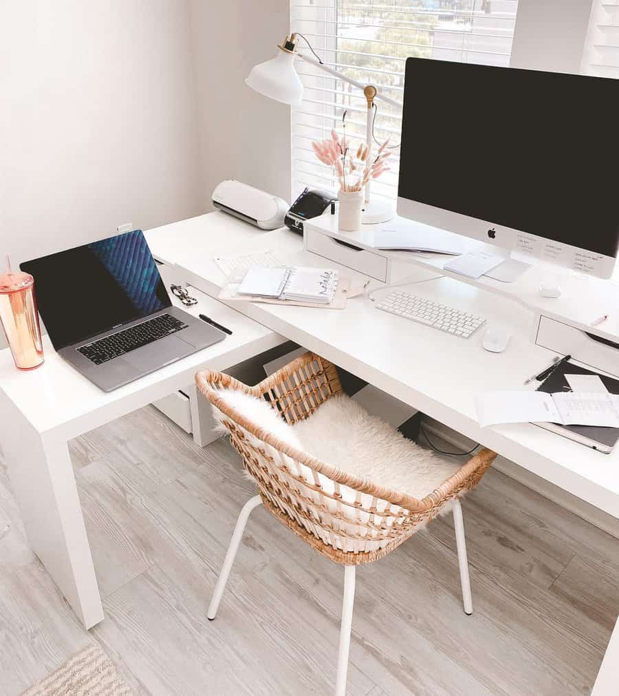 Minimalist home office with a white desk, computer, laptop, notebooks, a pink tumbler, plants, and a woven chair with a cushion