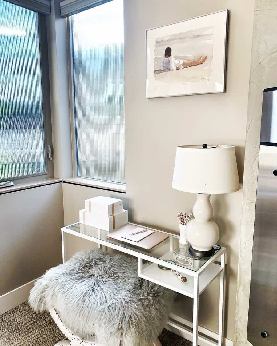 Modern nook with a glass desk, lamp, framed art, gift boxes, and a fur-covered stool by a window in neutral tones and natural light