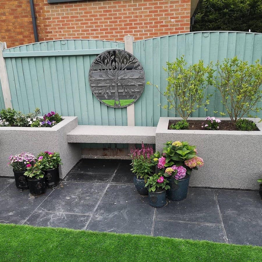 Garden corner with a concrete bench, flower pots, and a decorative metal wall art on a light blue fence