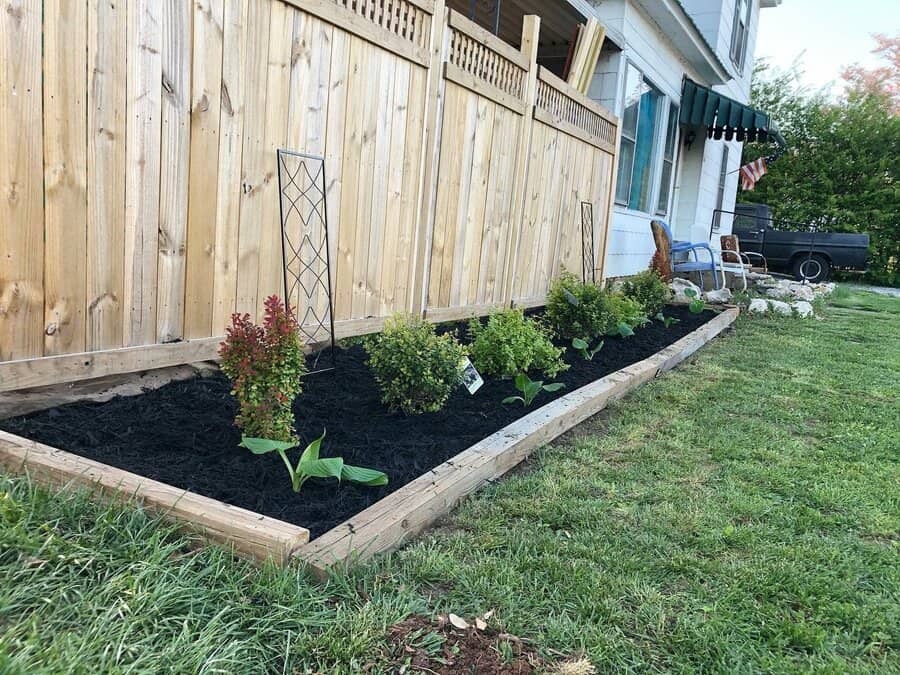 Garden bed with green plants and shrubs beside a wooden fence and building, bordered by grass