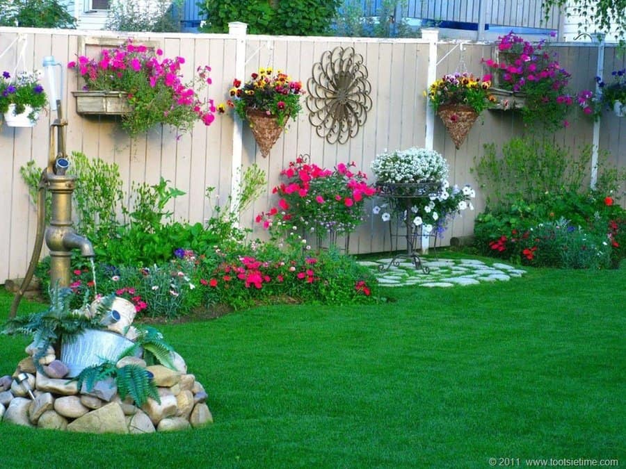 Lush garden with a wooden fence adorned with hanging flower baskets, metal wall art, a rustic water fountain, and colorful blooms