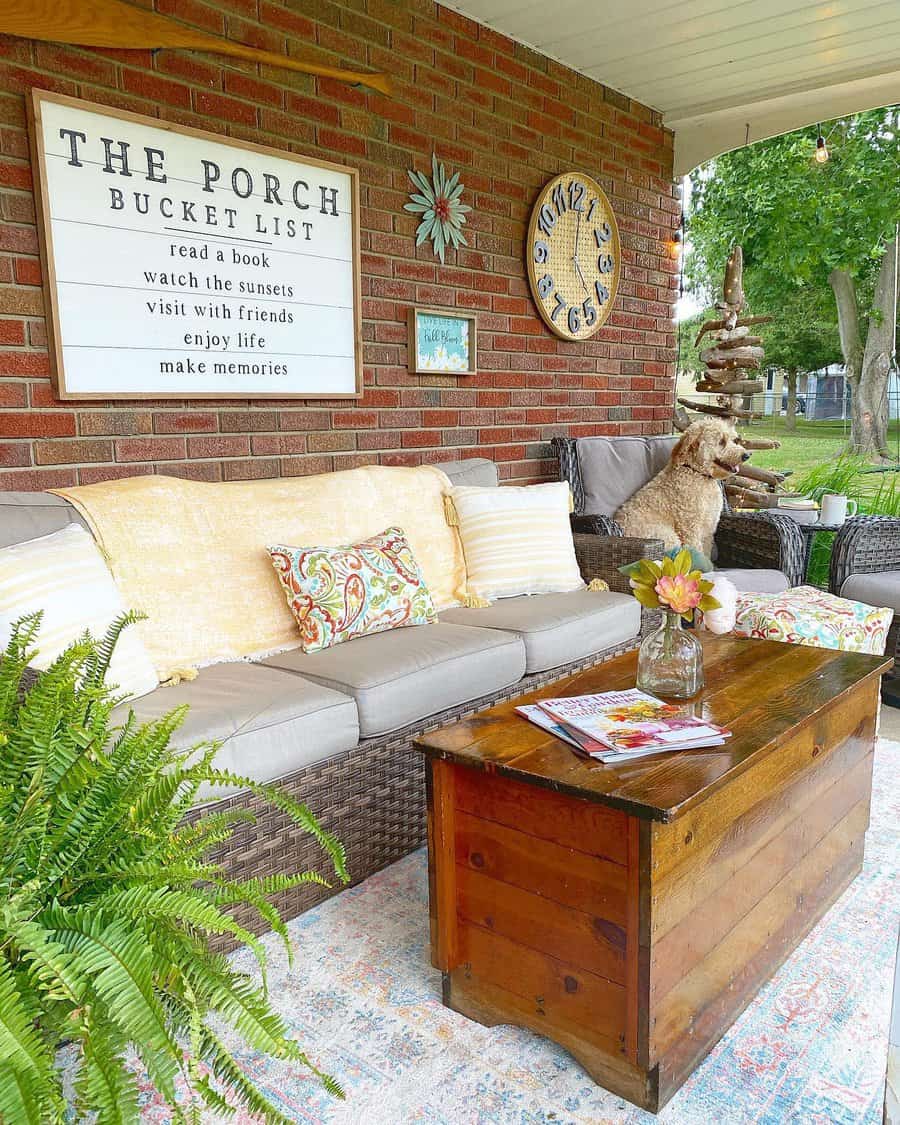 Cozy porch with wicker couch, fluffy dog, wooden table, plants, and a "Porch Bucket List" sign on brick wall