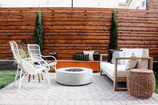 Outdoor patio with wicker chairs, cushioned sofa, a round fire pit on a tiled floor, and a wooden fence with tall plants