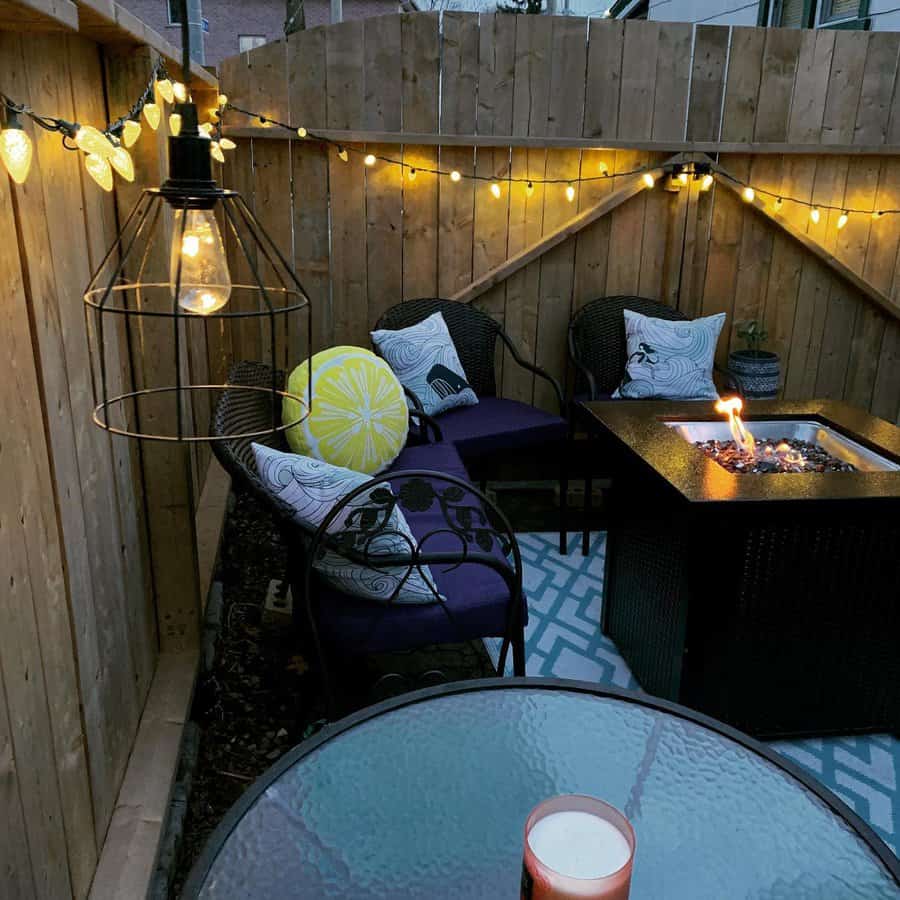 Cozy patio with string lights, wicker chairs with cushions, a lemon pillow, and a lit fire pit on a patterned rug