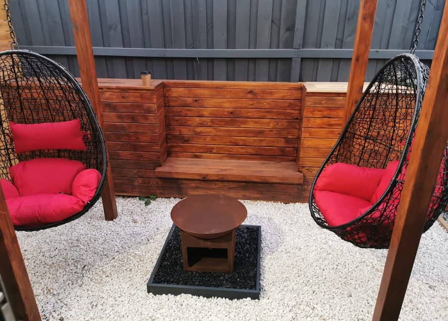 Two black hanging chairs with red cushions face a wooden bench. A round fire pit is centered on white gravel, enclosed by a dark fence