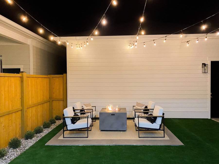 Outdoor patio with a fire pit surrounded by four cushioned chairs on a concrete slab, with string lights hanging overhead