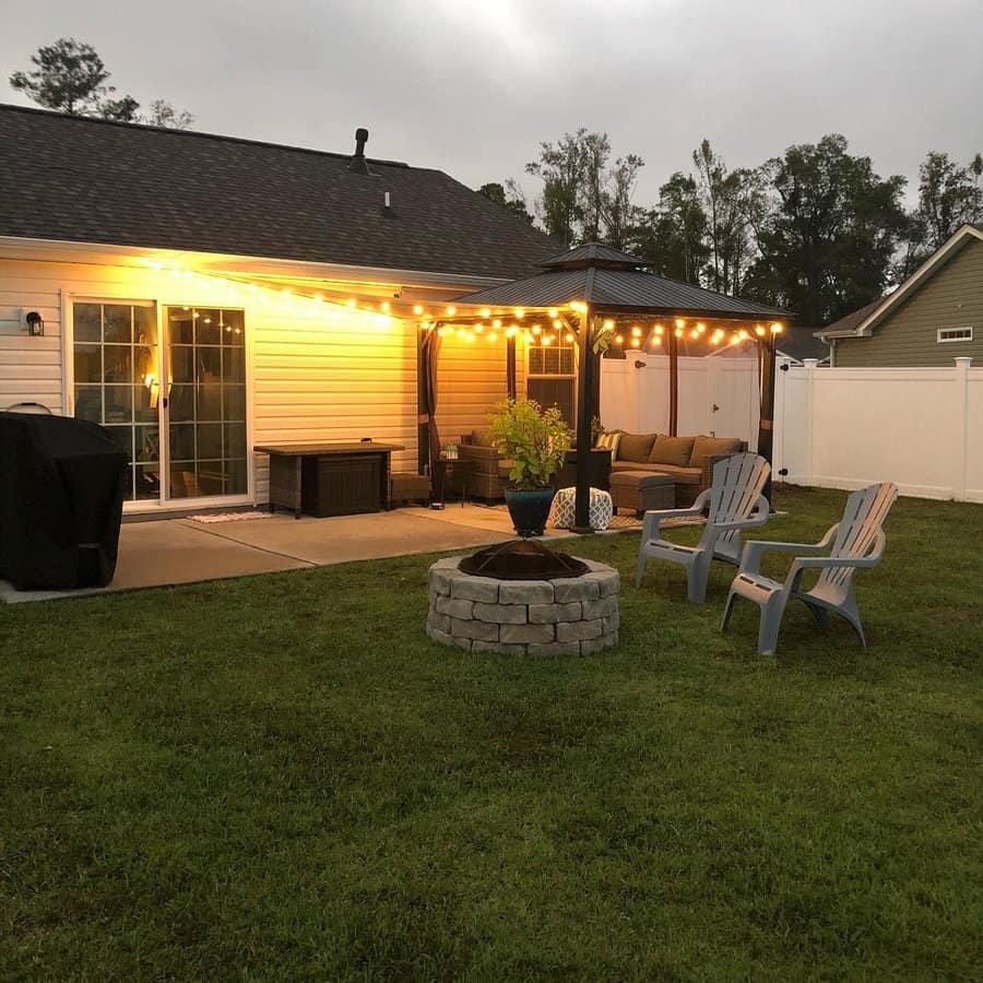 Backyard patio with string lights, outdoor furniture, fire pit, and lawn chairs on grass; house with sliding glass door at dusk