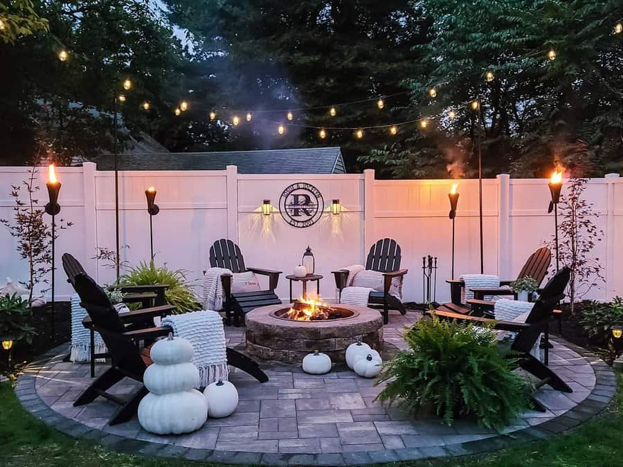 Cozy outdoor patio with a circular fire pit, Adirondack chairs, string lights, torches, pumpkins, and ferns, surrounded by a white fence