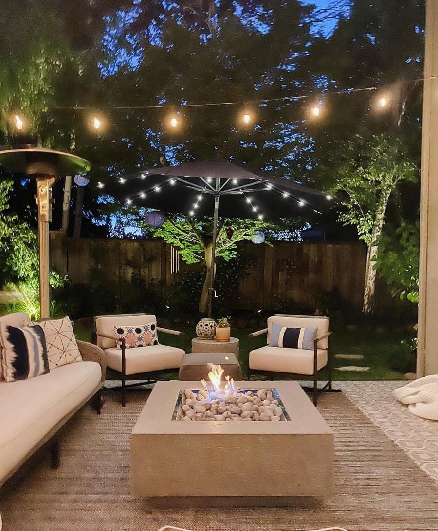 Cozy patio with armchairs and a sofa around a lit fire pit, string lights above, and an illuminated umbrella at dusk