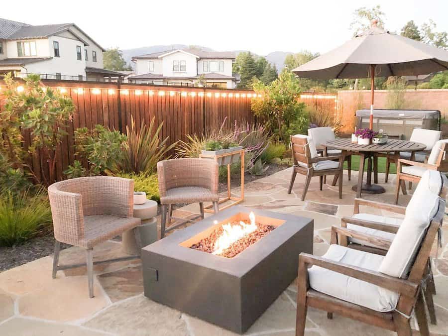 Outdoor patio with a fire pit, cushioned chairs, dining table with umbrella, and string lights on fence, surrounded by plants