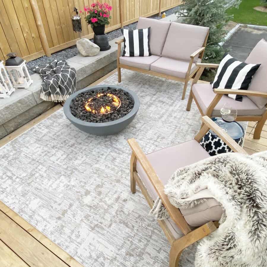 Cozy outdoor patio with beige furniture, a fire pit, striped pillows, and a soft rug; lanterns and blankets add warmth