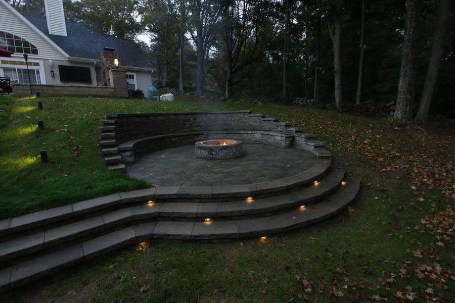 Outdoor fire pit area with circular stone seating, layered steps, built-in lighting, and a cozy backyard setting near a house