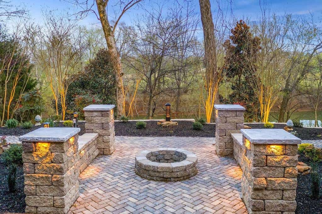 Elegant fire pit area with stone benches, warm lighting, herringbone brick patio, and a scenic view of trees and a river