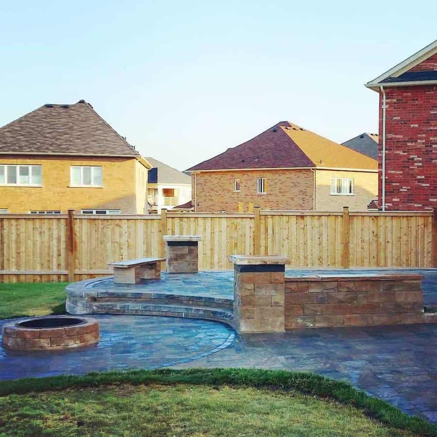 Simple backyard fire pit with stone seating, a paved patio, wooden privacy fence, and suburban neighborhood view