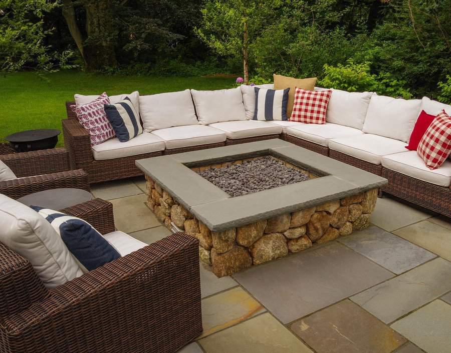 Outdoor patio with wicker furniture, red and white cushions, surrounding a stone fire pit on a tiled floor, set in a green garden