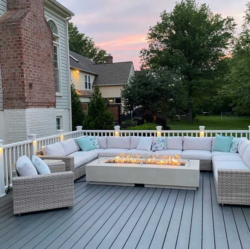 Outdoor deck with a large L-shaped sofa and a fire pit table, surrounded by trees, with a house in the background at sunset